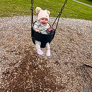 Cecelia is registered to the contest to win money with this photo: baby, child, swing, playground, hat, pink_hat, grass, wood_chips, outdoor, park, happy, smiling, clothing, footwear, person, fun, play, seat, chain, nature