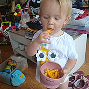 Innaya a rejoint le concours — aidez-le/la à gagner de superbes lots ! toddler, child, snack, cheese_puffs, pink_bowl, white_shirt, flower_design, socks, toys, plastic_container, folded_clothes, shoes, indoor, floor, cabinet, doll, colorful_toy, person, hair, face