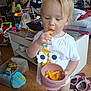 toddler, child, snack, cheese_puffs, pink_bowl, white_shirt, flower_design, socks, toys, plastic_container, folded_clothes, shoes, indoor, floor, cabinet, doll, colorful_toy, person, hair, face
