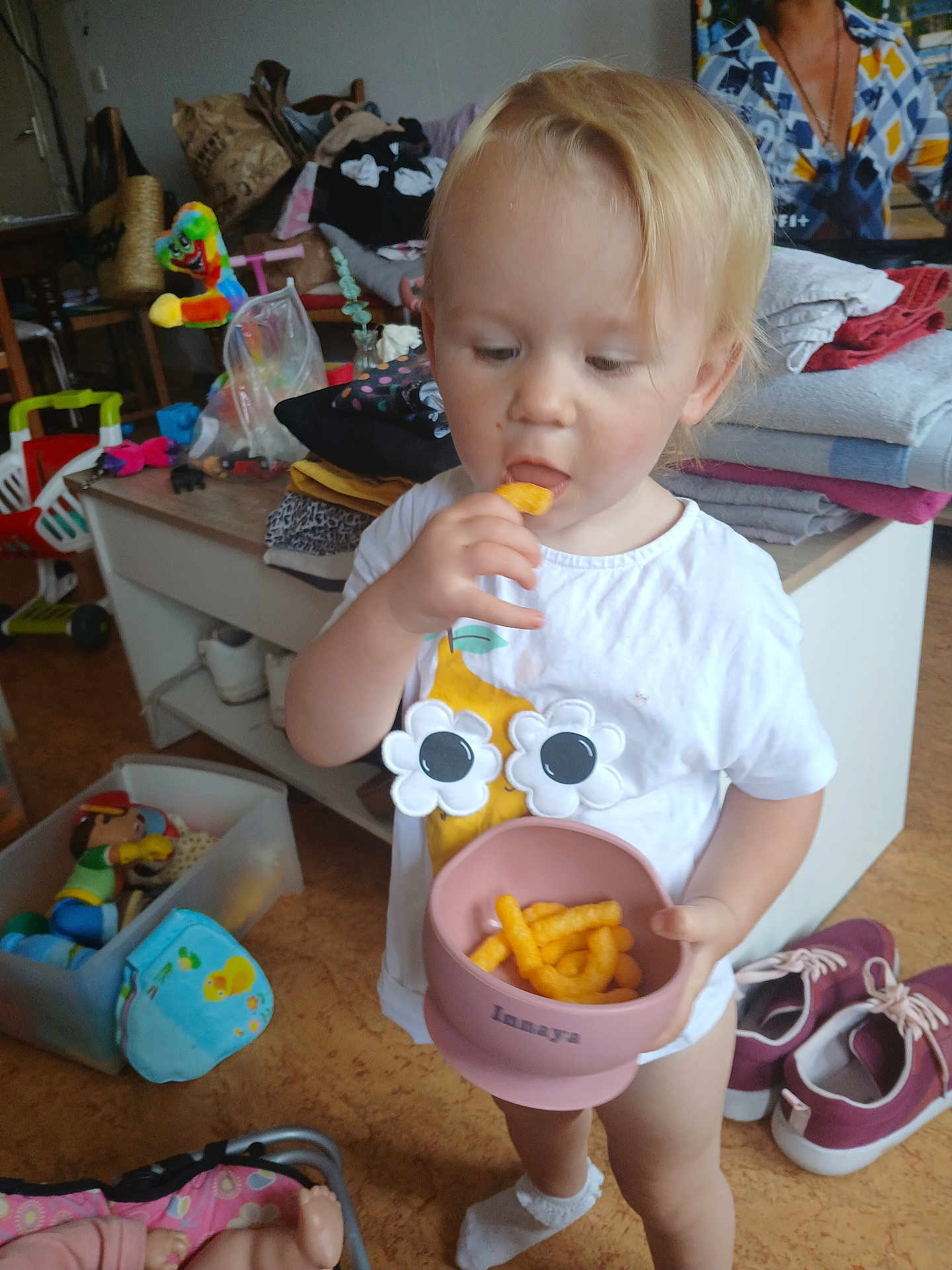 Innaya a rejoint le concours — aidez-le/la à gagner de superbes lots ! toddler, child, snack, cheese_puffs, pink_bowl, white_shirt, flower_design, socks, toys, plastic_container, folded_clothes, shoes, indoor, floor, cabinet, doll, colorful_toy, person, hair, face