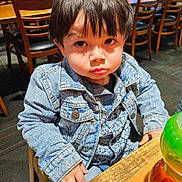 Maximus is registered to the contest to win money with this photo: child, toddler, denim_jacket, patterned_shirt, table, wooden_chair, restaurant, indoor, toy, serious_expression, face, hair, person, clothing, seated, furniture, casual, young_child, curious, portrait