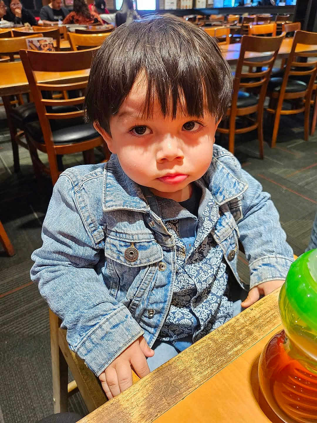Maximus is registered to the contest to win money with this photo: child, toddler, denim_jacket, patterned_shirt, table, wooden_chair, restaurant, indoor, toy, serious_expression, face, hair, person, clothing, seated, furniture, casual, young_child, curious, portrait