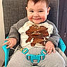 Axel is registered to the contest to win money with this photo: baby, child, high_chair, food, crumbs, smile, cute, clothing, shirt, pants, seatbelt, indoor, person, happy, messy, face, hands, hair, expression, furniture