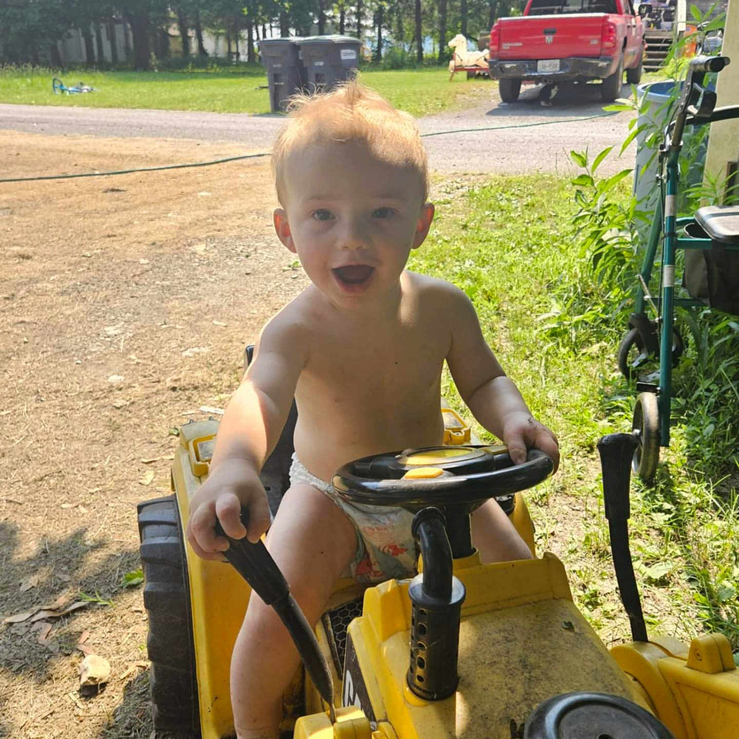 Oakley joined the competition — help win amazing prizes! toddler, child, toy_tractor, outdoor, sunlight, grass, tree, red_truck, steering_wheel, barefoot, happy, smiling, summer, diaper, play, nature, vehicle, yard, greenery, daytime