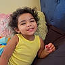 toddler, child, smiling, curly_hair, yellow_tank_top, bed, pillow, headboard, smartphone, social_media, indoor, happy, person, casual_clothing, relaxed, furniture, technology, portrait, home, cute