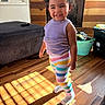 child, couch, curly_hair, fireplace, happy, home, indoors, laundry_basket, living_room, portrait, purple_top, rainbow_colors, smiling, socks, standing, striped_clothing, sunlight, toddler, window, wooden_floor