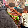 candid_photo, casual_clothing, children, curly_hair, doll, family, green_chair, hair_styling, home, indoor, living_room, playing, pretend_play, siblings, smiling, sofa, sunlight, toddler, toy, windows