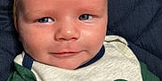 Abel is registered to the contest to win money with this photo: baby, infant, face, smile, clothing, green_shirt, white_shirt, hair, skin, eyes, portrait, closeup, cute, child, young, person, resting, quilt, fabric, background