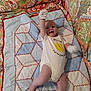 baby, infant, onesie, mittens, quilt, patterned_blanket, smiling_sun, text_on_clothing, raising_arm, lying_down, colorful, fabric, cute, happy, child, indoors, soft_texture, comfort, playful, portrait