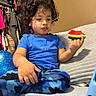 child, toddler, watermelon, blue_shirt, blue_pants, camouflage, curly_hair, indoor, couch, blanket, food, snack, sitting, hand, face, expression, furniture, room, casual_clothing, relaxed