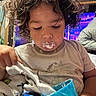 toddler, child, curly_hair, eating, spoon, cup, messy, indoors, clothing, face, portrait, focused, food, hand, blanket, cozy, cute, young_child, mess, casual