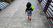 Roberto is registered to the contest to win money with this photo: back_view, ball, bridge, casual_clothing, child, concrete, curly_hair, daytime, footwear, greenery, nature, outdoor, park, pedestrian_bridge, person, railings, shorts, trees, tshirt, walking
