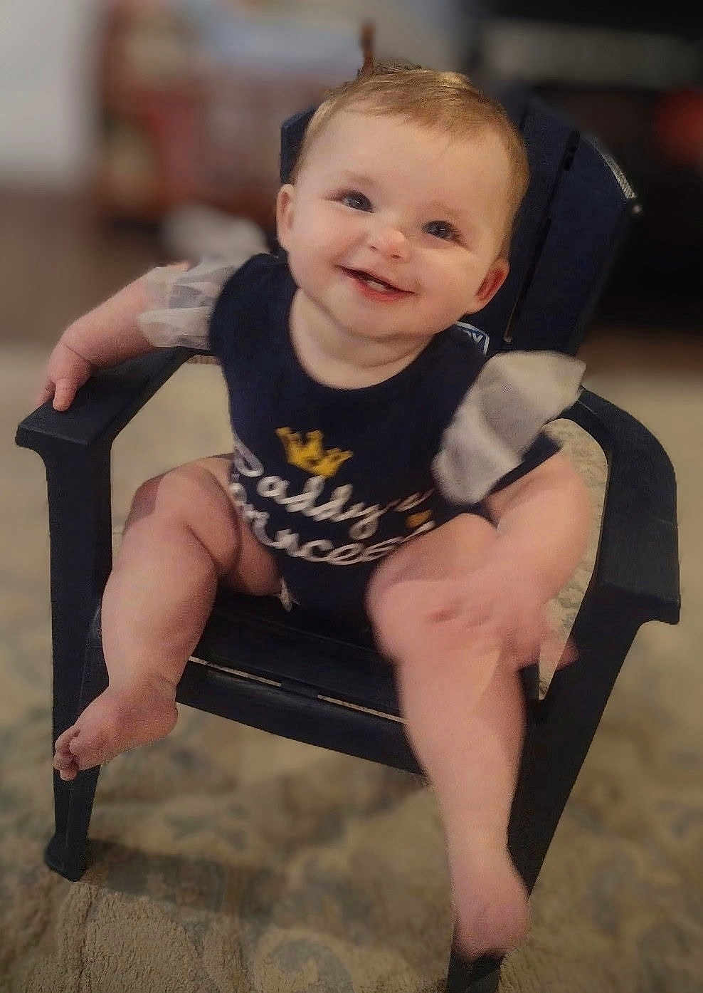Charlotte Elishka is registered to the contest to win money with this photo: baby, child, smile, happy, sitting, chair, clothing, indoor, cute, person, infant, puffy_sleeves, navy_outfit, text_on_clothing, footwear_free, legs, face, head, furniture, carpet
