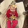 baby, baby_rocker, bow, child, comfort, cushion, cute, expression, flooring, hand, headband, indoor, infant, person, plush_toy, red_clothing, seat_belt, small, toy, wood_floor