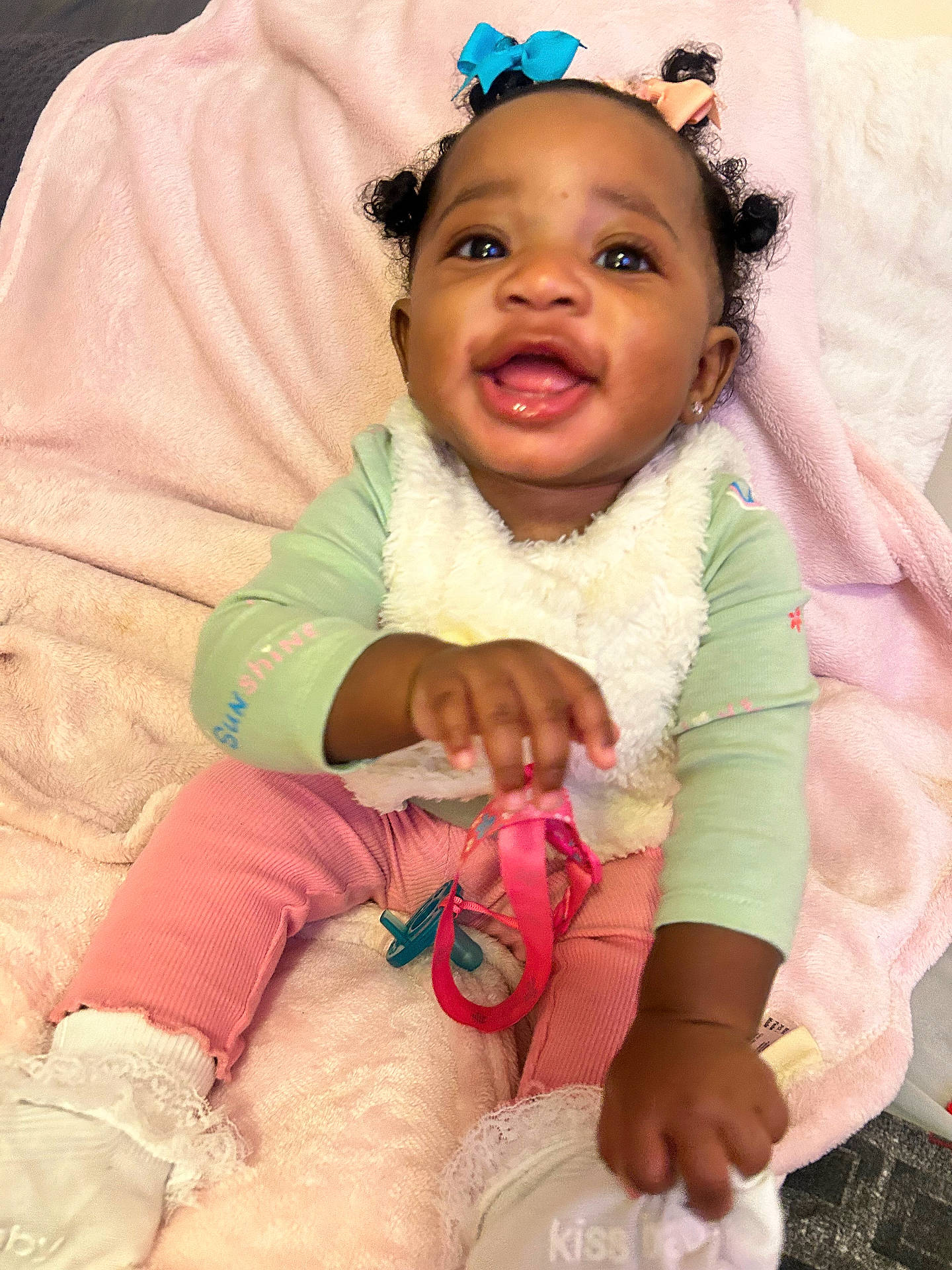 A Niylah joined the competition — help win amazing prizes! baby, baby_toy, blanket, bows, child, curly_hair, green_shirt, hair_accessories, happy, indoor, infant, lace, pastel_clothing, pink_blanket, pink_pants, portrait, sitting, smiling, toy, white_socks