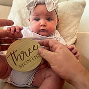 Oakley is registered to the contest to win money with this photo: baby, infant, headband, bow, plaque, wooden, three_months, hands, couch, pillow, clothing, face, legs, skin, person, indoors, sitting, cute, portrait, child