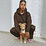 woman, dog, puppy, brown_hoodie, braided_hair, hoop_earrings, pink_slippers, white_brick_wall, outdoor, sunlight, sidewalk, casual_clothing, young_adult, pet, crouching, portrait, urban, fashion, calm, daylight