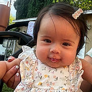 Dashly is registered to the contest to win money with this photo: adult_hand, baby, backyard, brown_eyes, cheeks, child, close_up, dark_hair, face, family, floral_dress, garden, hair_bow, infant, outdoor, portrait, smiling, stroller, sunlight, trees