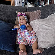 Dashly is registered to the contest to win money with this photo: baby, child, infant, smiling, happy, portrait, indoor, couch, sofa, pillows, dress, floral_dress, headband, bow, rattle, toy, sitting, barefoot, home, furniture