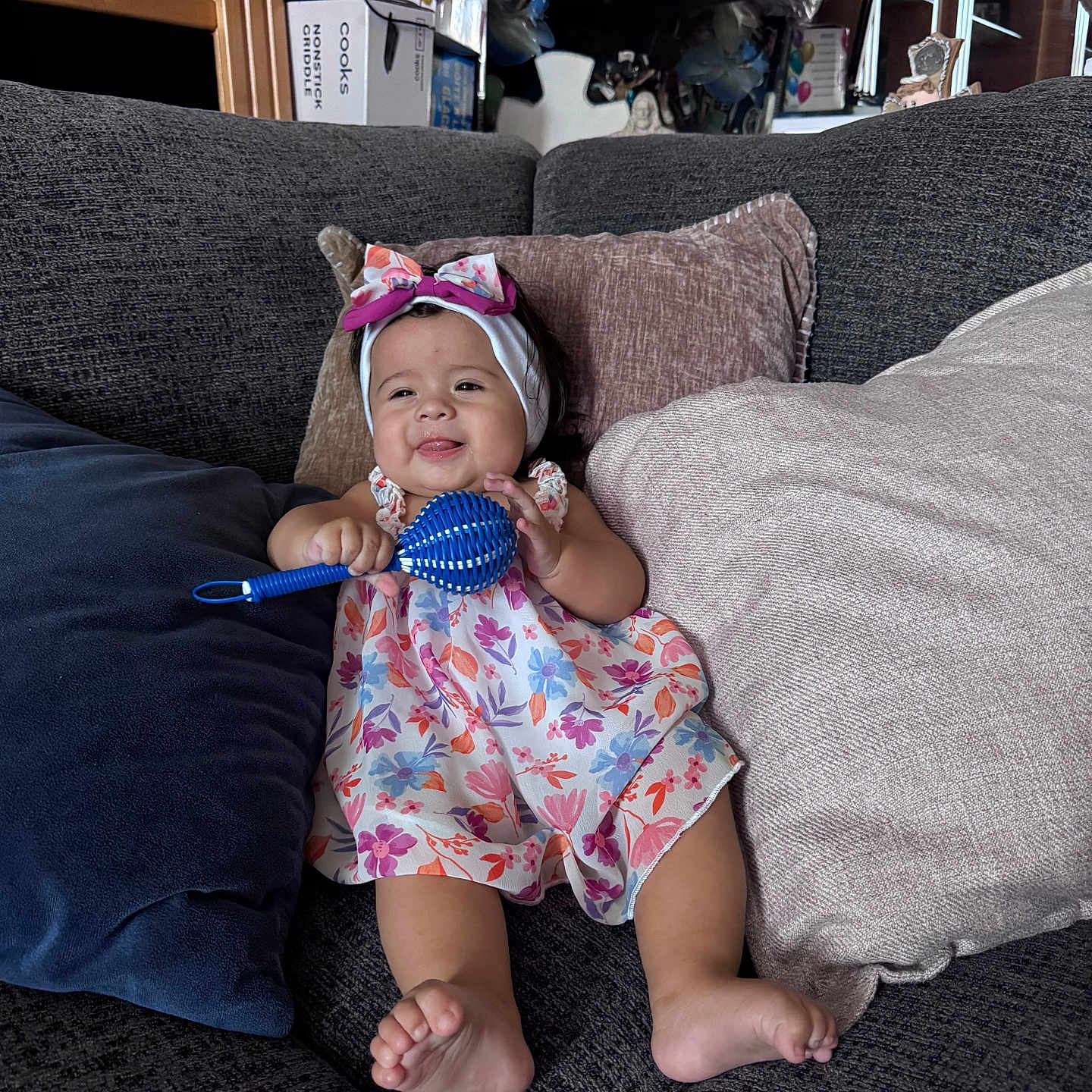 Dashly is registered to the contest to win money with this photo: baby, child, infant, smiling, happy, portrait, indoor, couch, sofa, pillows, dress, floral_dress, headband, bow, rattle, toy, sitting, barefoot, home, furniture