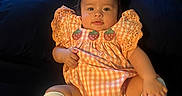 Dashly is registered to the contest to win money with this photo: baby, bed, black_hair, checkered_dress, child, cozy, dark_background, dress, eyes, face, fashion, hair_clip, hands, indoors, infant, knee_socks, legs, portrait, sitting, strawberry_pattern