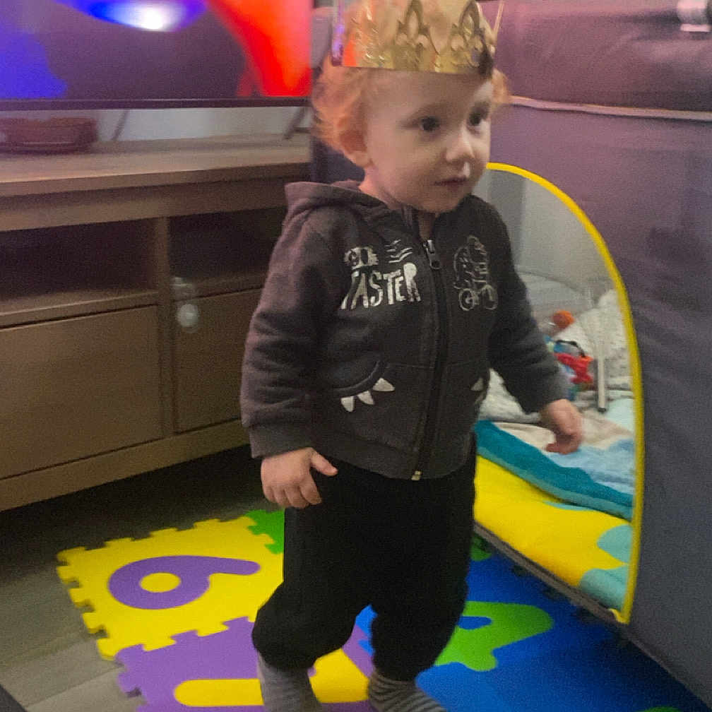 Tyméo participe au concours pour gagner de l'argent avec cette photo : toddler, child, crown, foam_mat, numbers, indoor, living_room, television, vacuum_cleaner, clothing, hoodie, socks, floor, furniture, playtime, curious, young_child, home, toy, colorful
