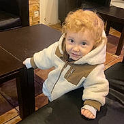 Tyméo participe au concours pour gagner de l'argent avec cette photo : toddler, child, curly_hair, fleece_jacket, table, bench, indoor, floor_tiles, brown_furniture, looking_up, smiling, cozy_clothing, young_child, person, casual, curious, warm_lighting, hands, standing, portrait