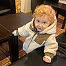 toddler, child, curly_hair, fleece_jacket, table, bench, indoor, floor_tiles, brown_furniture, looking_up, smiling, cozy_clothing, young_child, person, casual, curious, warm_lighting, hands, standing, portrait