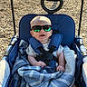 baby, sunglasses, stroller, blanket, outdoor, sunlight, child, smile, happy, seatbelt, blue, clothing, footwear, person, infant, playground, woodchips, shade, reflection, comfort