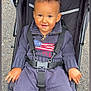 american_flag, baby, casual, child, clothing, daylight, face, footwear, hands, happy, head, jogging_pants, outdoor, pavement, person, portrait, seatbelt, stroller, sweater, toddler
