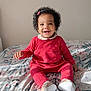 baby, toddler, child, smiling, curly_hair, hair_bow, red_outfit, onesie, sitting, bed, bedding, floral_pattern, white_shoes, portrait, indoor, happy, cute, skin, eyes, hands