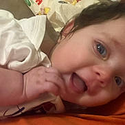 Remington is registered to the contest to win money with this photo: baby, child, face, blue_eyes, white_clothing, smiling, hand, infant, closeup, lying_down, soft_skin, happy, cheeks, cute, portrait, playful, indoors, orange_surface, young, person