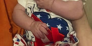 Remington joined the competition — help win amazing prizes! baby, child, infant, patriotic_clothing, dress, stars, stripes, hand, fingers, sitting, person, soft_surface, orange, cute, indoor, holding, calm, curious, face, portrait