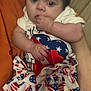 baby, child, infant, patriotic_clothing, dress, stars, stripes, hand, fingers, sitting, person, soft_surface, orange, cute, indoor, holding, calm, curious, face, portrait