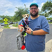 Nikki is registered to the contest to win money with this photo: bag, blue_shirt, cap, casual_clothing, clouds, crosswalk_sign, dog, glasses, happy_dog, leash, man, medal, outdoor, parking_lot, pavement, sidewalk, sky, smiling, street, trees