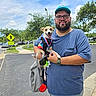 man, dog, outdoor, sidewalk, street, trees, medal, glasses, cap, blue_shirt, smiling, happy_dog, leash, bag, pavement, sky, clouds, crosswalk_sign, parking_lot, casual_clothing
