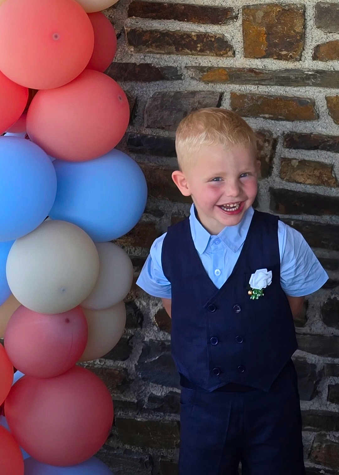 Enzo participe au concours pour gagner de l'argent avec cette photo : child, boy, smiling, balloons, vest, shirt, stone_wall, blue, red, pastel_colors, happy, portrait, party, celebration, flower_boutonniere, short_hair, indoors, cute, formal_wear, standing
