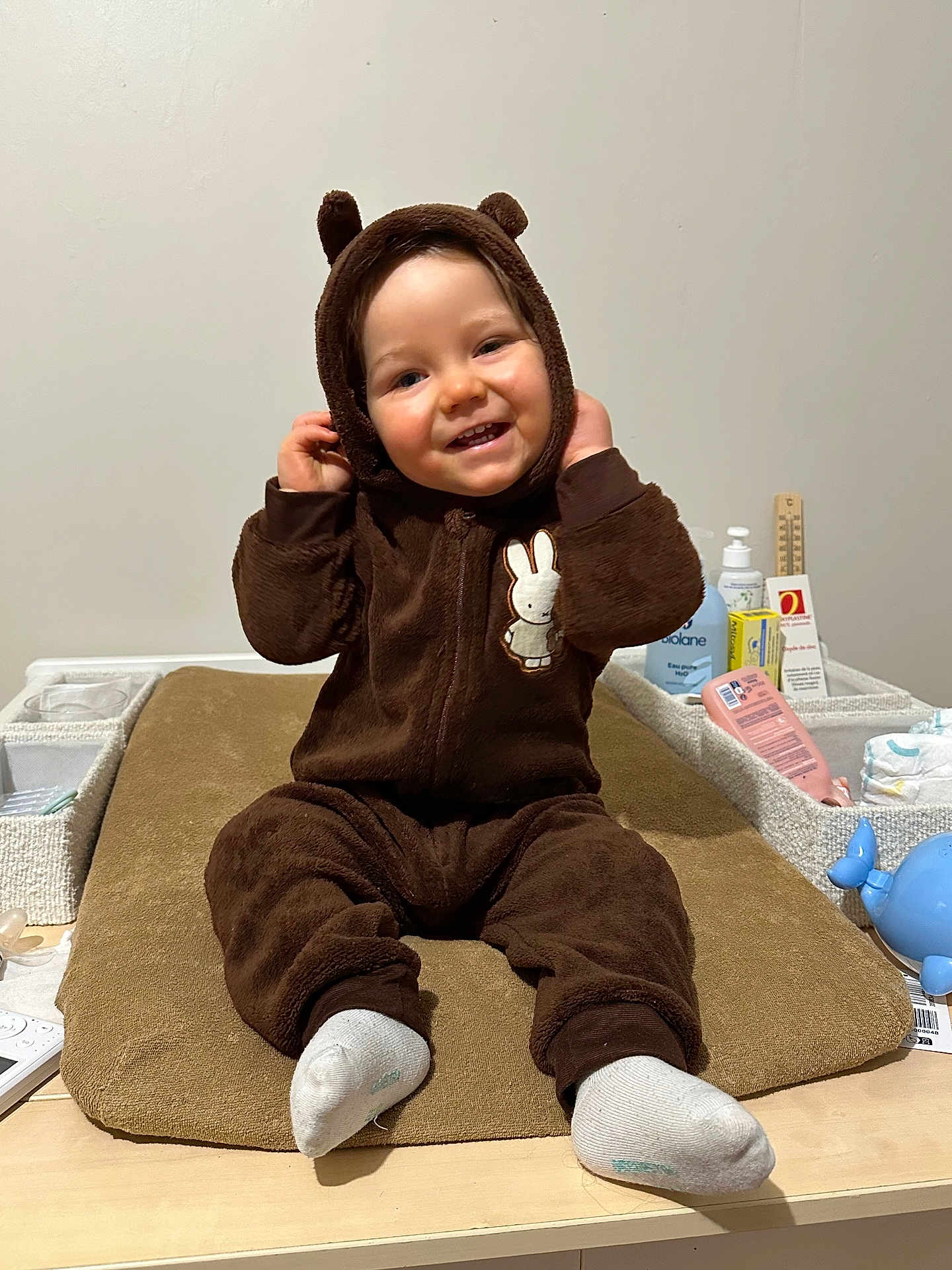 Elyo a rejoint le concours — aidez-le/la à gagner de superbes lots ! toddler, child, baby, onesie, brown_clothing, smiling, face, hood, ears, sitting, changing_pad, baby_care, bottle, diapers, table, indoor, cute, happy, person, infant