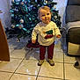 blanket, child, christmas_tree, couch, cute, decorations, festive, floor_tiles, furniture, holiday, indoors, lighting, living_room, ornaments, pants, person, shoes, smiling, sweater, toddler