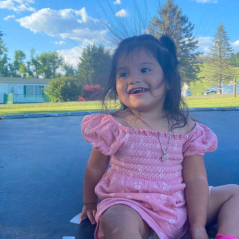 Esperanza is registered to the contest to win money with this photo: blue_sky, casual, child, clouds, girl, grass, hair, happy, necklace, outdoor, pink_dress, playful, shoes, smiling, static_electricity, sunny_day, text_overlay, toddler, trampoline, trees