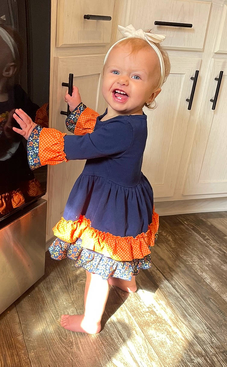 Kacie is registered to the contest to win money with this photo: baby_toddler_clothing, child, day_dress, door, electric_blue, finger, flooring, fun, gesture, happy, hardwood, human_leg, joint, one_piece_garment, orange, person, sleeve, smile, thumb, toddler