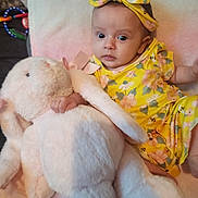 Alivia is registered to the contest to win money with this photo: baby, infant, stuffed_animal, plush_toy, yellow_clothing, headband, flower_pattern, bed, blanket, cute, child, soft_toy, indoor, portrait, young_child, baby_girl, toy, comfort, resting, colorful