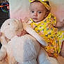 baby, infant, stuffed_animal, plush_toy, yellow_clothing, headband, flower_pattern, bed, blanket, cute, child, soft_toy, indoor, portrait, young_child, baby_girl, toy, comfort, resting, colorful