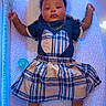baby, infant, playpen, plaid_skirt, heart_socks, blue_lighting, pacifier, cute_clothing, child, lying_down, soft_toys, mesh_sides, patterned_blanket, adorable, portrait, indoors, calm_expression, small_hands, feet_up, necklace