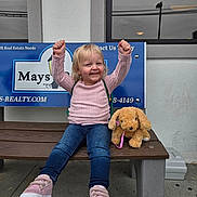 Harper is registered to the contest to win money with this photo: toddler, child, bench, pink_sweater, jeans, sneakers, plush_toy, dog_toy, smile, happy, outdoor, real_estate_sign, window, reflection, concrete, building_wall, raised_arms, casual_clothing, daylight, portrait