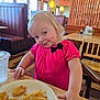 toddler, child, restaurant, food, plate, nuggets, french_fries, pink_shirt, black_bow, table, cup, water, wooden_chair, booth_seating, window, light_fixture, smile, cute, indoor, dining