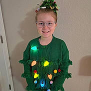 Adaline joined the competition — help win amazing prizes! child, glasses, smile, green_sweater, christmas_lights, pom_poms, holiday_headpiece, indoor, wall, carpet, power_outlet, door, festive, happy, person, portrait, long_sleeves, standing, light_up, casual