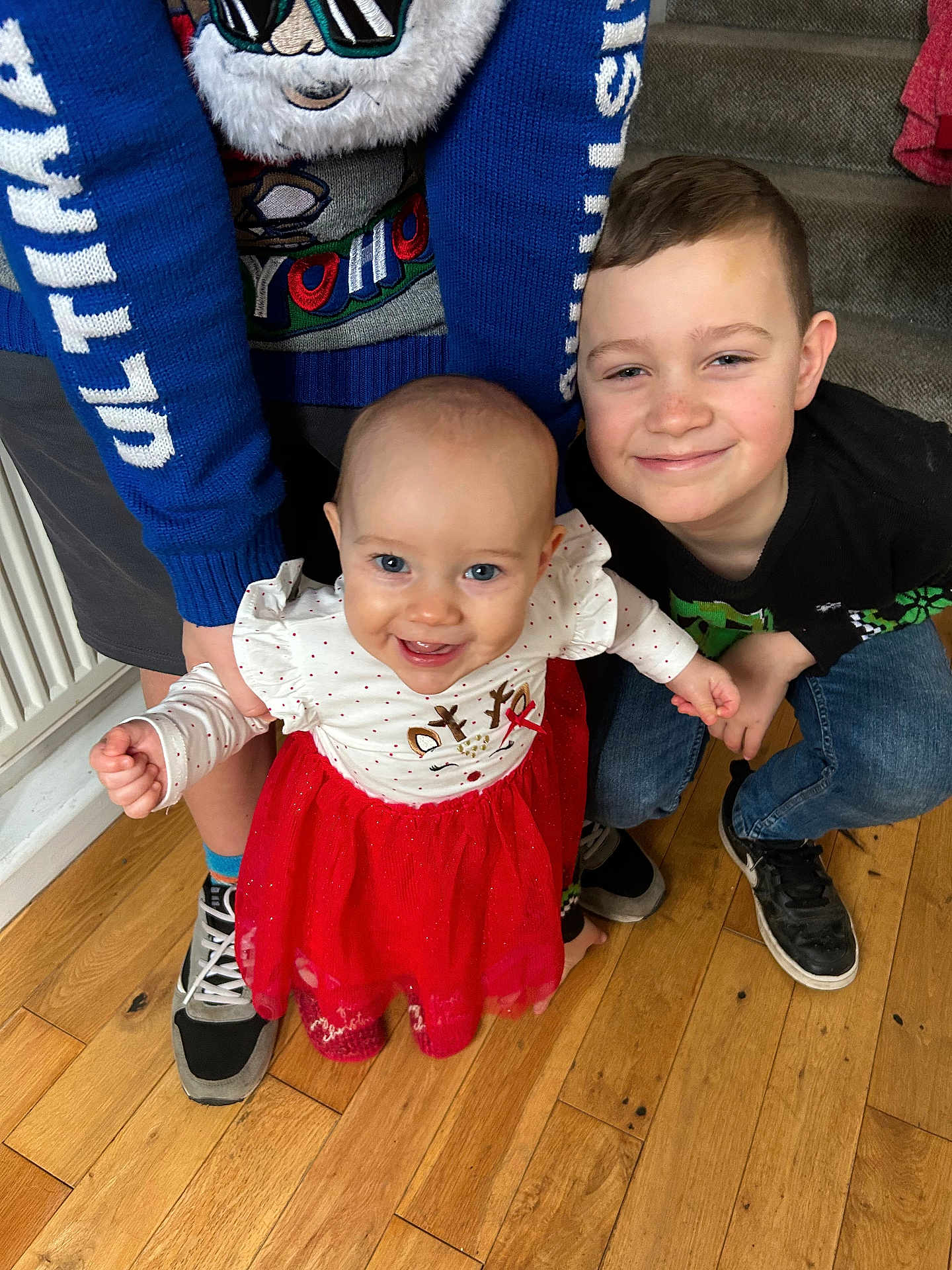 Edie joined the competition — help win amazing prizes! baby, child, smiling, red_dress, blue_sweater, indoor, wooden_floor, holding_hands, crouching, happy, people, footwear, radiator, clothing, boy, person, family, portrait, cute, toddler