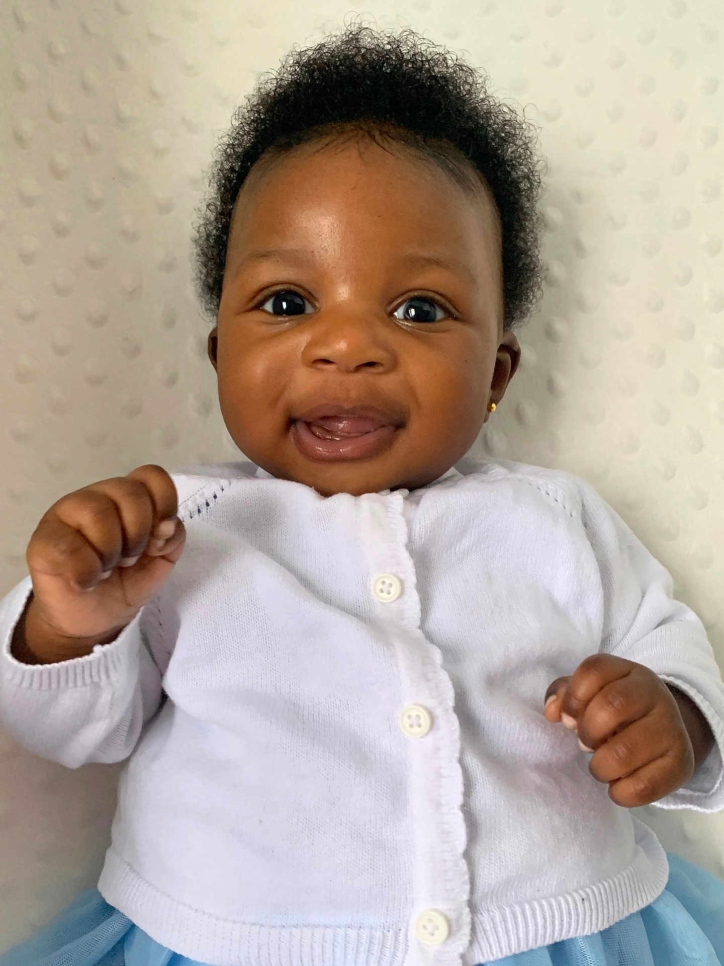 Momoririoluwa joined the competition — help win amazing prizes! baby, smiling, face, hands, white_sweater, blue_skirt, curly_hair, earring, portrait, closeup, happy, cute, child, infant, indoor, soft_background, buttoned_clothing, skin, fingers, expression