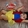 Aitana is registered to the contest to win money with this photo: baby, child, costume, dress, yellow_skirt, red_bow, blue_top, blanket, soft_texture, face, arms_up, curly_hair, lying_down, decorative_sign, name_sign, cartoon_birds, indoor, portrait, cute, infant